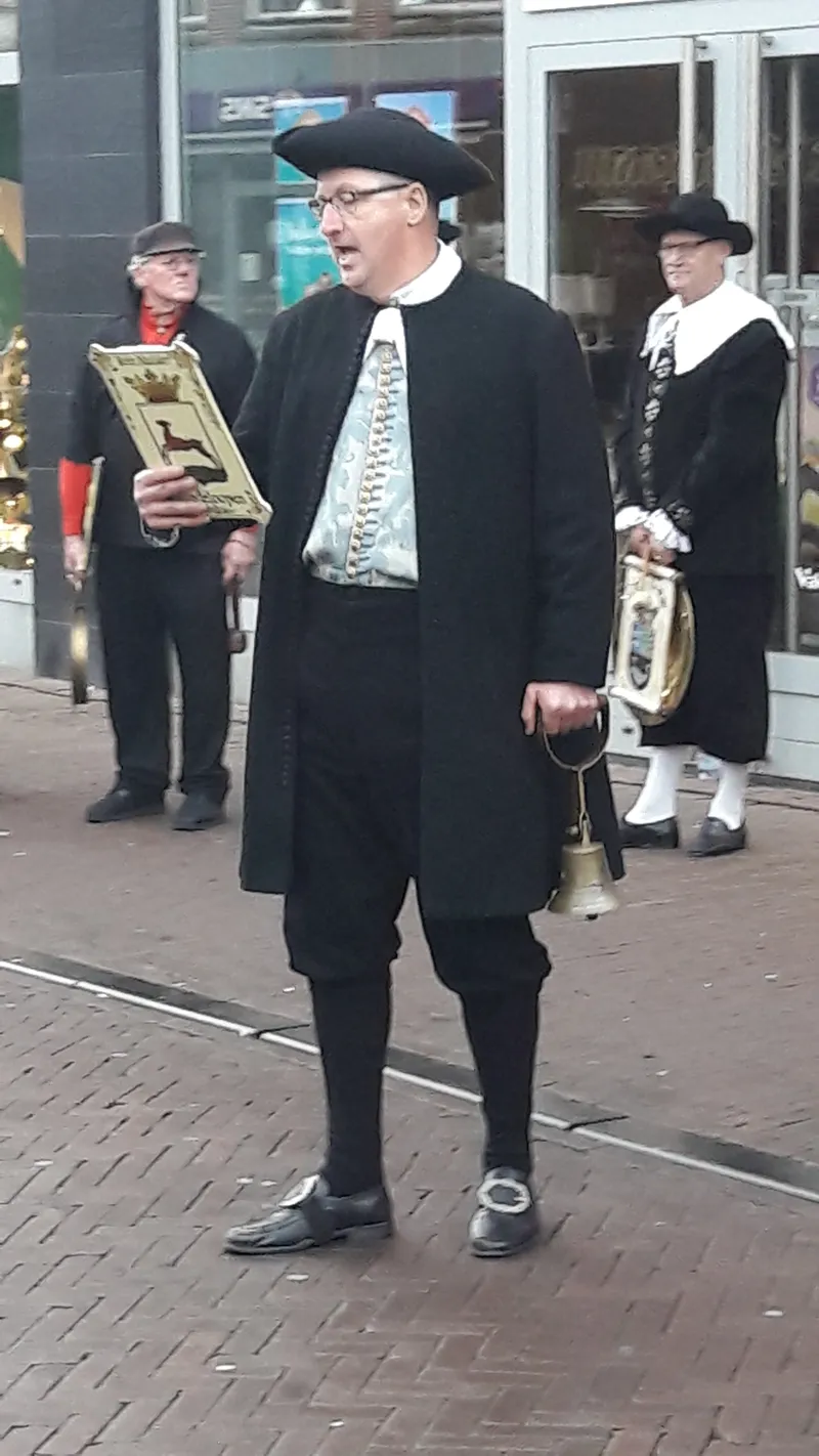Man in Hindelooper dracht met driekante hoed en bel leest voor van een beschilderd bord